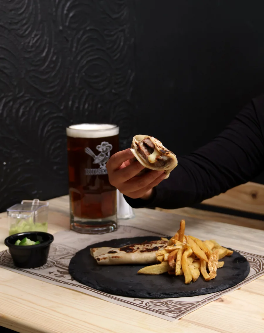 Burrito norteño sobre plato negro con papas fritas, junto a una cerveza obscura
